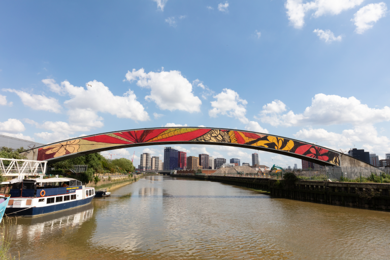 A bridge with organic curves and floral designs - red, yellow, blue, and white - spans across a river. Beneath its central arch, a skyline of buildings is visible in the distance. The sky is bright blue with scattered clouds, giving the scene a vibrant and airy feel.